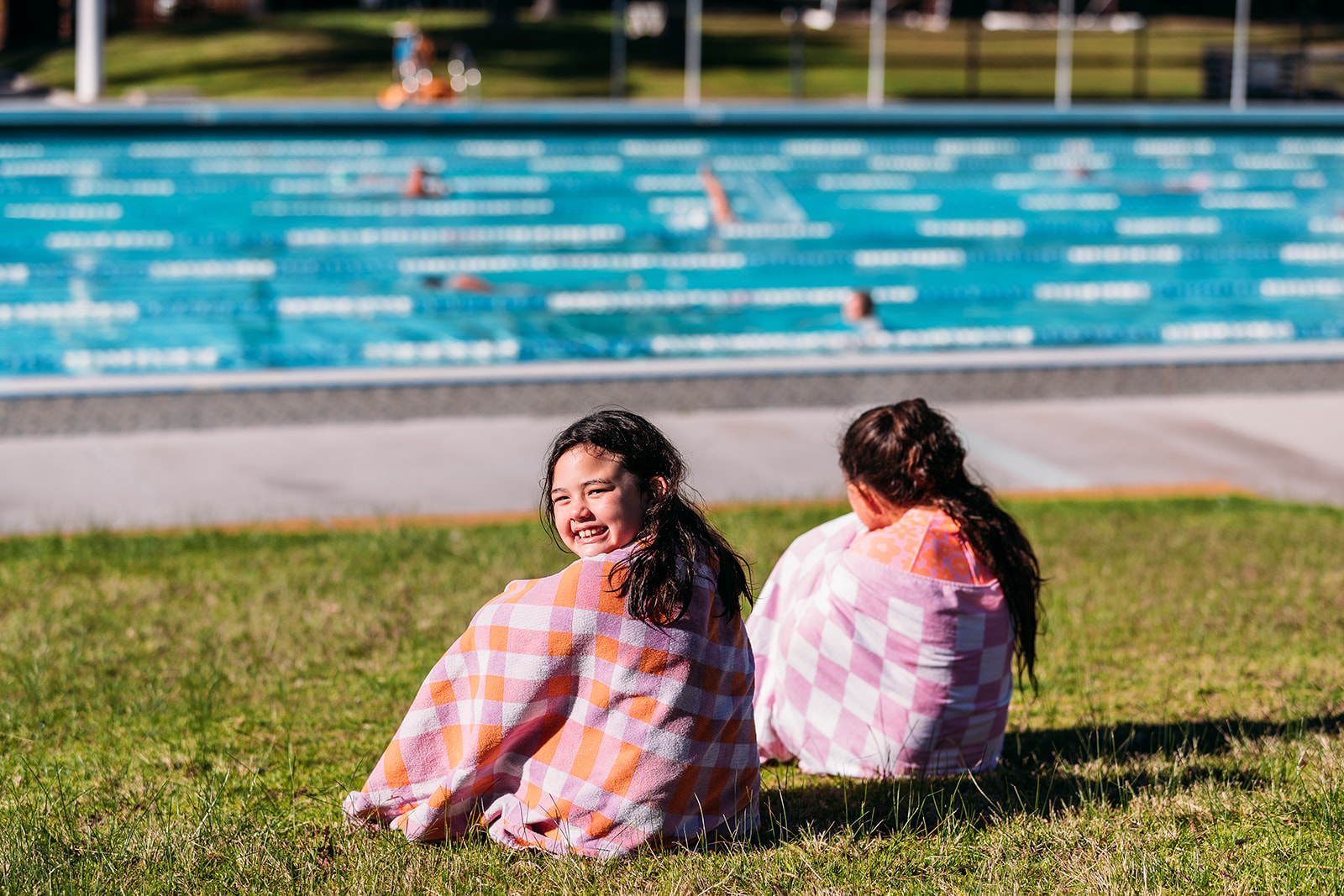 2 girls in towels sitting near pool looking at the camera.